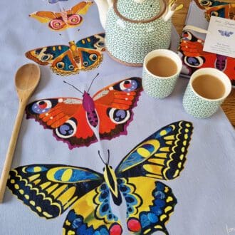 A pale purple tea towel, printed with five digitally designed butterflies in a line, with the smallest at the top and the largest at the bottom. The tea towel is laid on a table with a teapot and two small cups of tea to one side and a wooden spoon to the other.