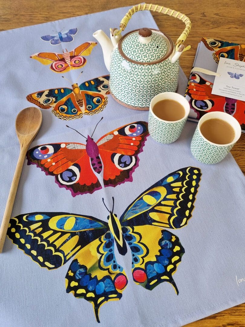 A pale purple tea towel, printed with five digitally designed butterflies in a line, with the smallest at the top and the largest at the bottom. The tea towel is laid on a table with a teapot and two small cups of tea to one side and a wooden spoon to the other.