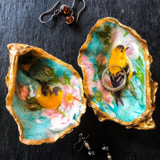 Pair of gilded decoupage oyster shell trinket dishes decorated with yellow song birds lie on a dark table surrounded by jewellery