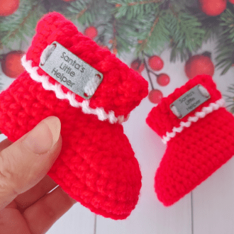 a pair of red Christmas Baby Booties with a white edging and an Ultrasuede tag saying 'Santa's Little Helper' available in sizes newborn, 0-3 and 3-6 months