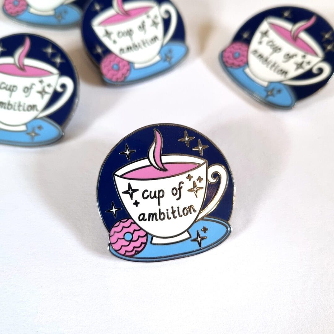 A close up of an enamel pin badge featuring a white tea cup with the words 'cup of ambition' in the centre. Around the cup there is a halo of navy blue with stars, and a pink biscuit on the side of the saucer the mug is sat on.
