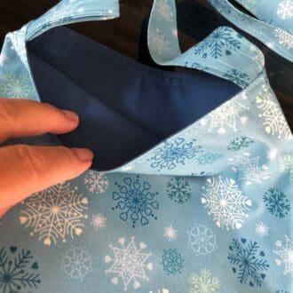Adjustable cross body style mastectomy drain bag in a pale blue fabric covered with blue and white snowflakes and a dusky mid blue cotton lining, on a reflective black surface
