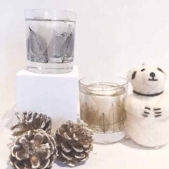 Pair of Winter Shades gel and soy wax candles, one in cool silver shades, the other in warm copper surrounded by fir cones and a polar bear tree decoration.