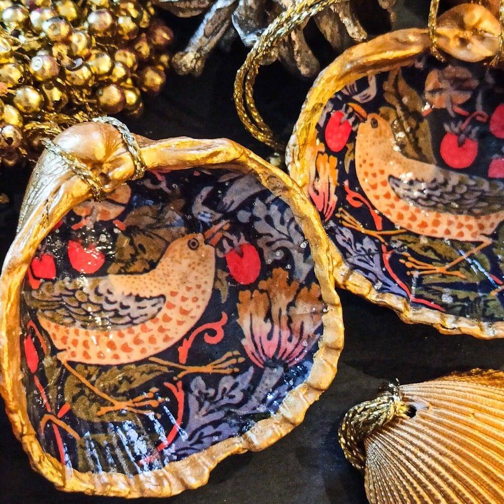 A pair of William Morris gilded, decoupaged, Strawberry Thief cockle shell Christmas decorations are surrounded by gold berries and a pine cone on a dark surface.