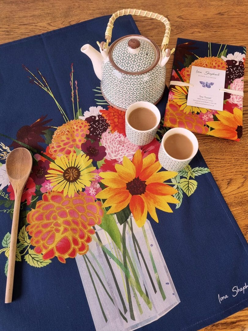 A navy blue tea towel printed with an illustration of a colourful mixed bouquet of flowers in a jam jar. The tea towel is laid out on the table with teapot and two small cups of tea to the right hand side.