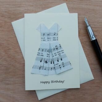 Happy Birthday cream coloured card lying flat on envelope showing origami folded dress made using sheet music. Writing says Happy Birthday. Is pictured with fountain pen next to it.