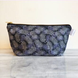 In the centre of the image is a blue cotton make up bag with a hand painted palm leaf print and finished with a brass zip across the top.