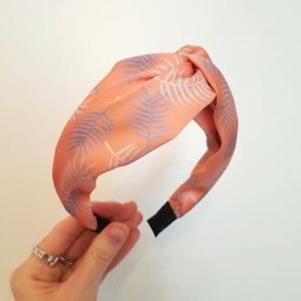 A hand is holding a pink headband at an angle in the centre of the image. The Alice band is made from pink faux silk with purple palm leaves and a twist in the centre.