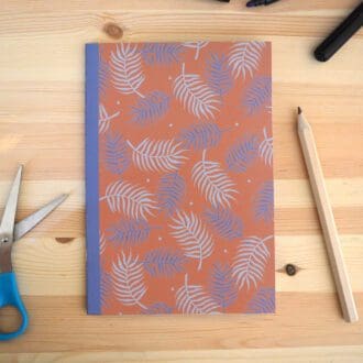 A pink notebook with purple palm leaf pattern is laid in the centre of a wooden desk with scissors and pencil and pen scattered around it.