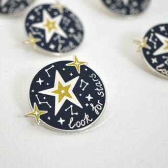 A close up of a round enamel pin badge with a dark blue background, silver and yellow stars, constellations and the words 'look for stars'.