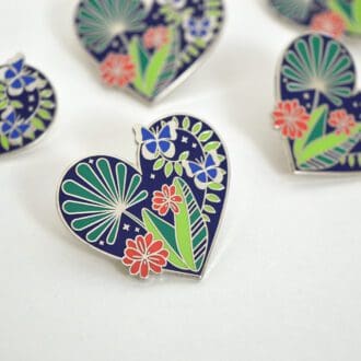 A close up of a heart shaped enamel pin badge with green rainforest foliage, orange flowers and two blue butterflies.