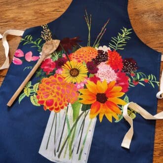 A navy blue apron printed with an illustration of a colourful mixed bouquet of flowers in a jam jar. The tea towel is laid out on the table with a wooden spoon down the left hand side