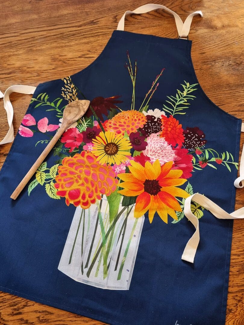 A navy blue apron printed with an illustration of a colourful mixed bouquet of flowers in a jam jar. The tea towel is laid out on the table with a wooden spoon down the left hand side
