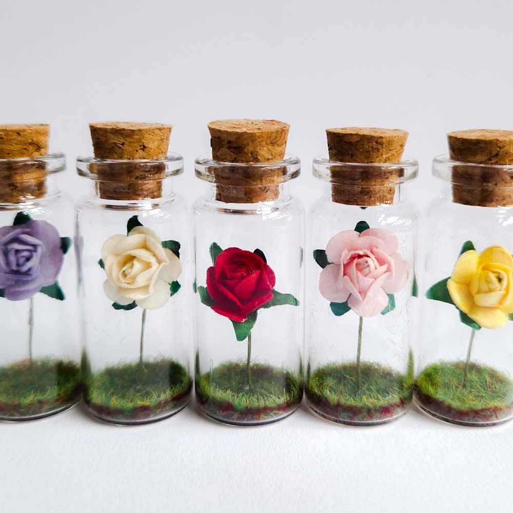 a row of miniature glass bottles containing different coloured paper roses, designed by under the blossom tree