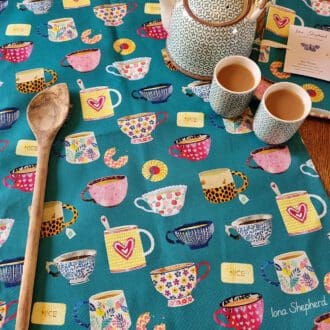 A teal coloured tea towel printed with illustrations of colourful tea cups and biscuits. The tea towel is laid on a table with a tea pot and two cups of tea to one side.