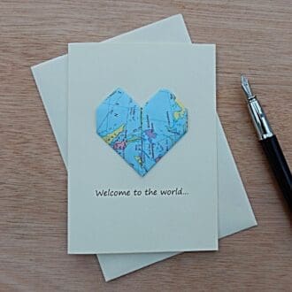 Cream coloured origami map heart new baby card lying on envelope next to a fountain pen. Card says Welcome to the World. Map heart shows countries of the world.