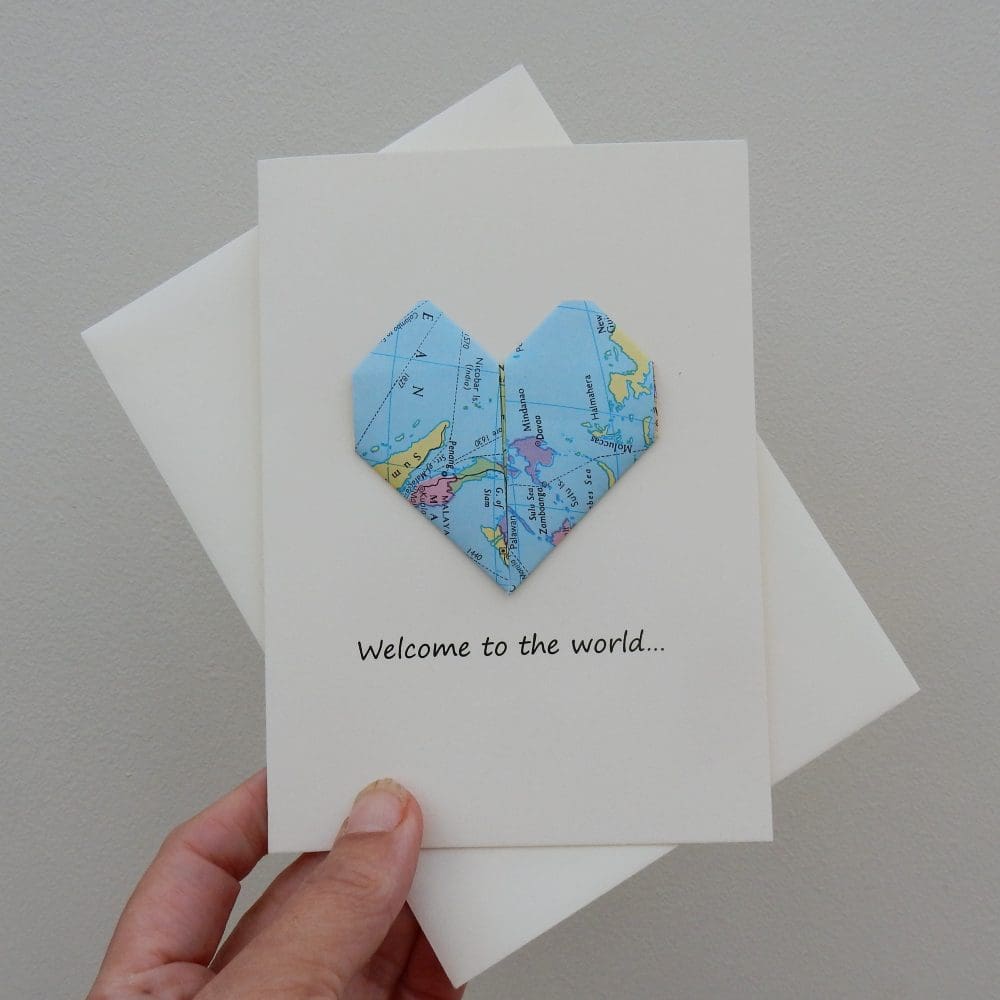 Hand holding Welcome to the World new baby card and envelope. Card has an origami heart made from an atlas page.