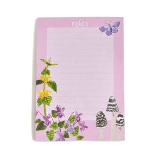 In the centre of the image is an A6, lined notepad illustrated with wild flora and fauna found in our British woodlands. The illustrations are set on a pink background.