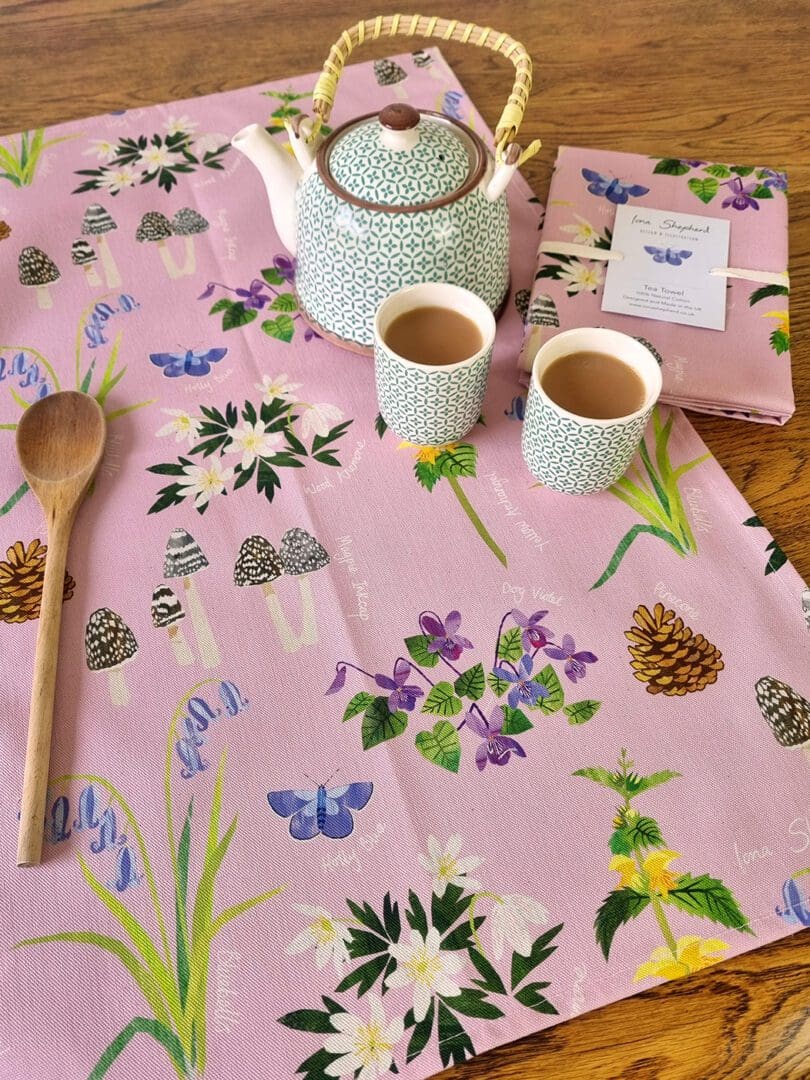 A pale pink tea towel, printed with digitally designed native woodland flowers and Holly Blue butterfly. The tea towel is laid on a table with a teapot and two small cups of tea to one side and a wooden spoon to the other.