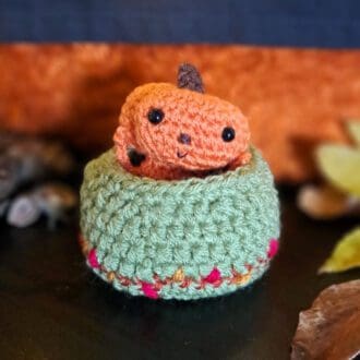 Crochet pumpkin sprite, an orange figure with a pumpkin for a head is sitting in a green embroidered nest and is waving at you!