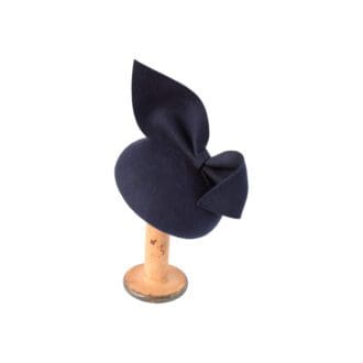 navy felt button hat with large bow trim perched on wooden stand on white background