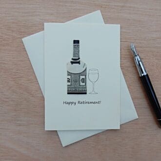 Happy retirement card lying flat on envelope with nearby fountain pen. Card shows origami bottle made from a sheet of monopoly money next to a wine glass. Cream coloured card with black and white bottle