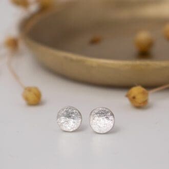 small round silver earrings rest on white background with small brass dish and dried flowers