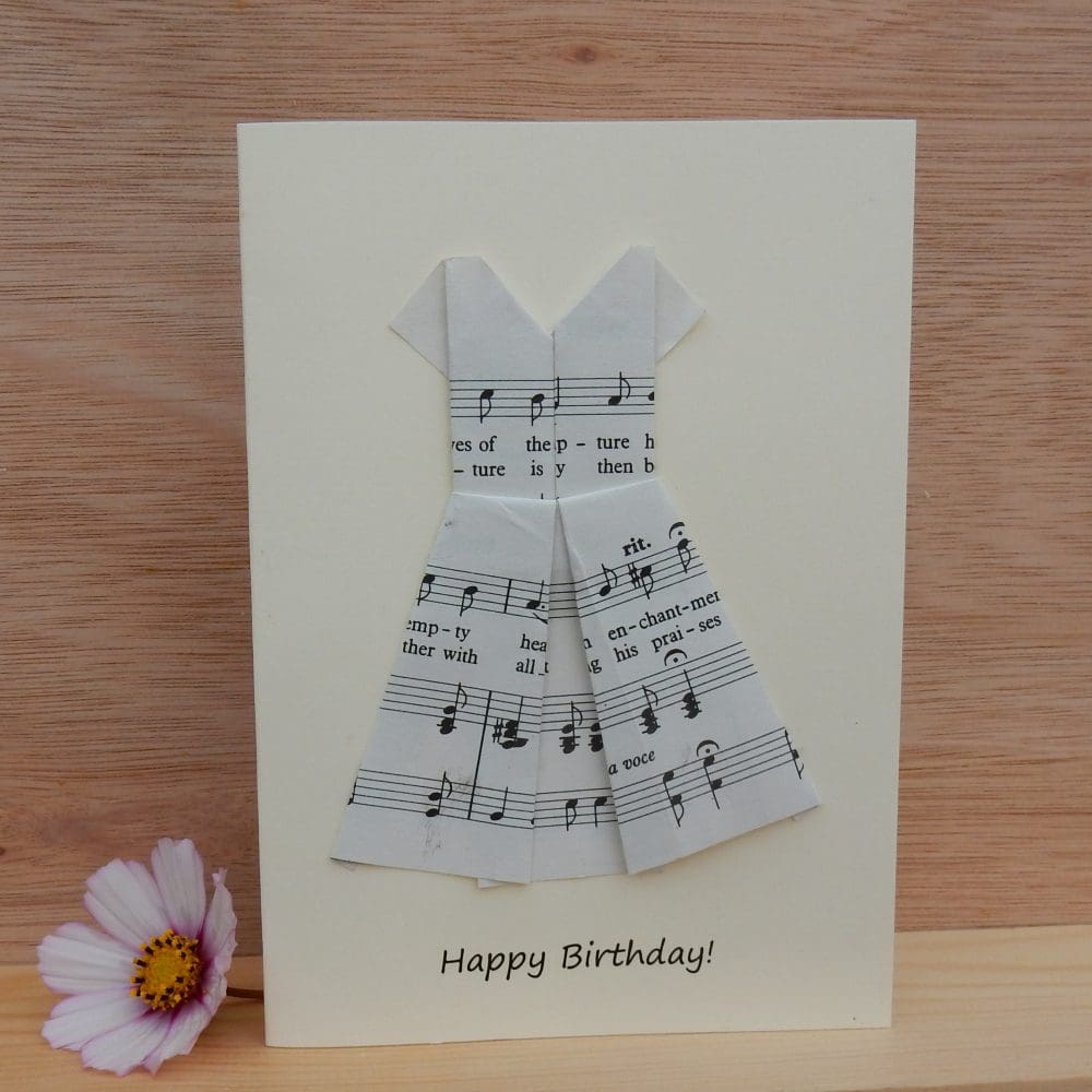 Happy Birthday card showing origami folded dress made using sheet music. Writing says Happy Birthday. Card is pictured standing on a shelf with a flower next to it.