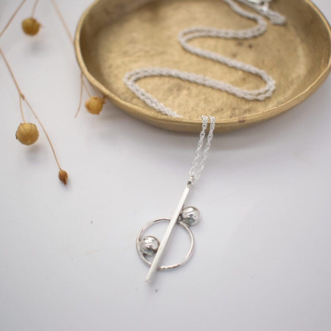 silver necklace on white background with small brass dish and dried flowers in background