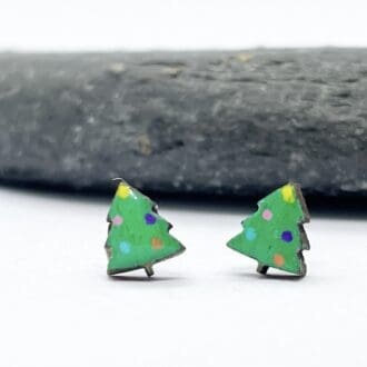 Green Christmas tree earrings that are decorated with multicoloured baubles on a white background in front of a grey slate stone