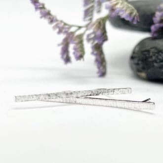 long silver bar earrings with a hammered texture on a white background with purple flowers and a slate stone