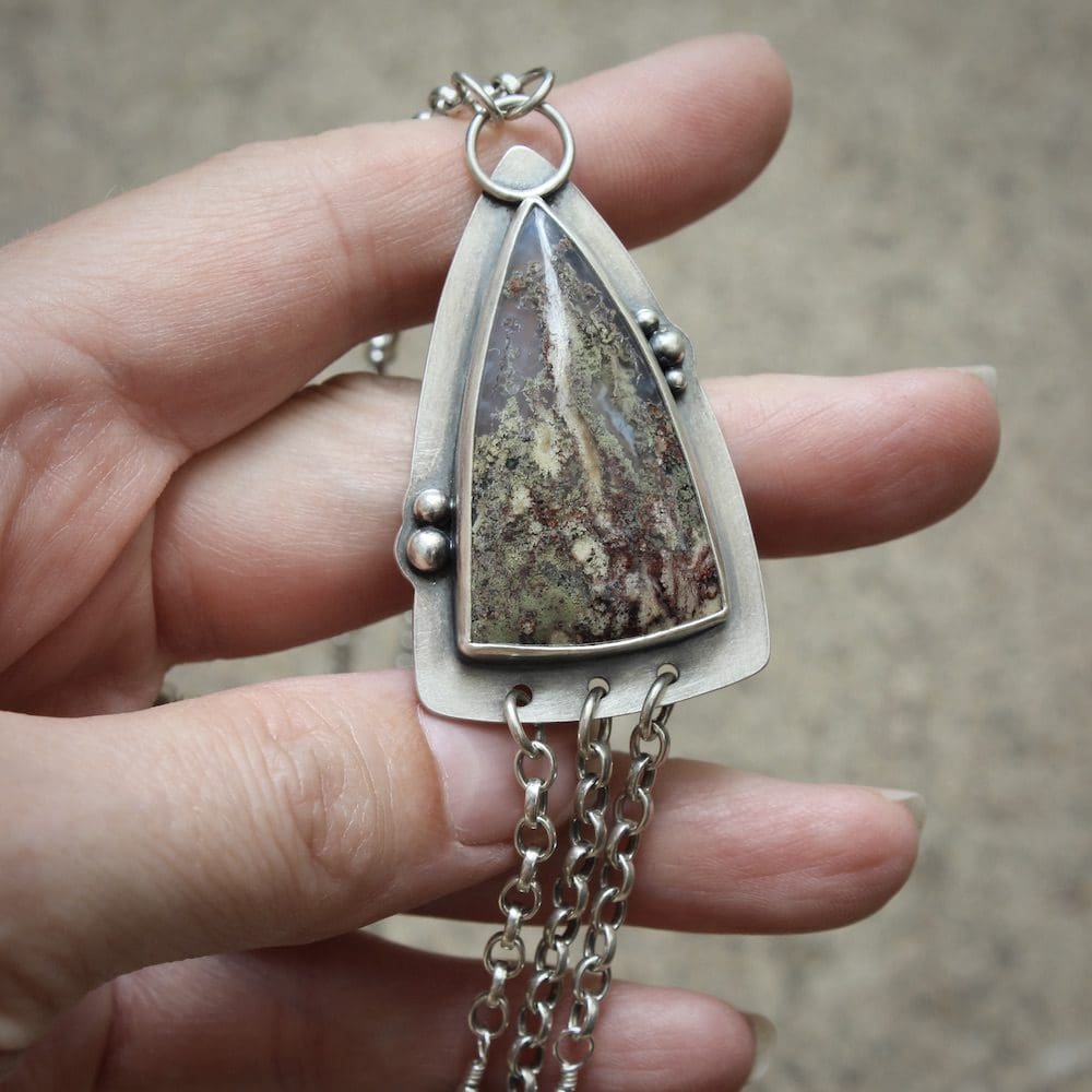 A sterling silver pendant shaped like an arrowhead, with a moss agate gemstone and round moss agate beads on a silver chain, held in the left hand of a woman, against a light brown concrete background.
