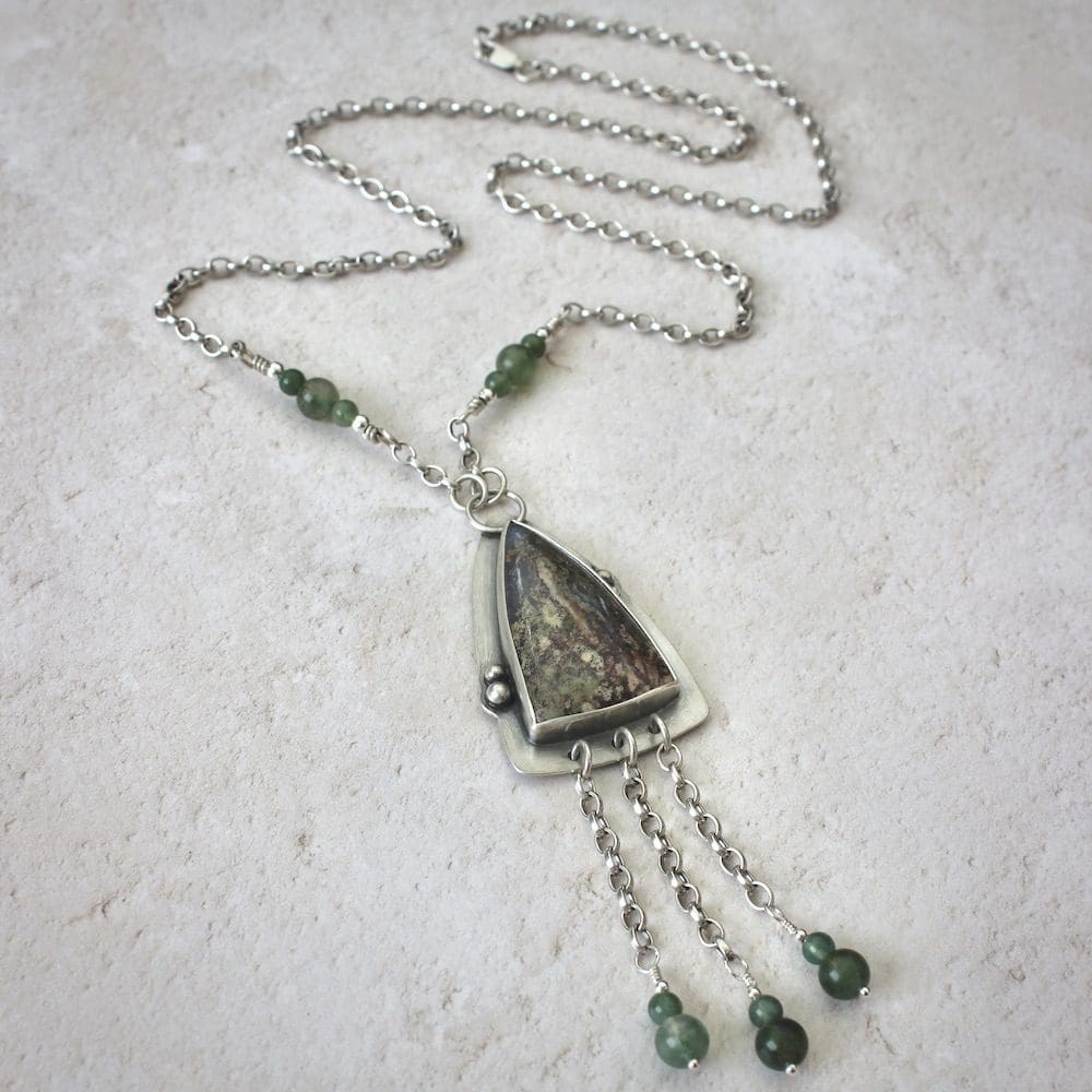 A sterling silver pendant shaped like an arrowhead, with a moss agate gemstone and round moss agate beads on a silver chain, on a white background.