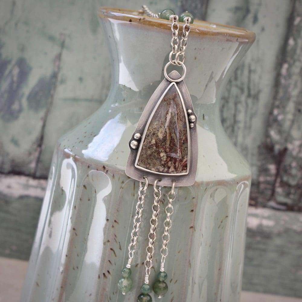 A sterling silver and moss agate gemstone pendant necklace with silver chain, laying on the neck of a green ceramic vase against a green background.