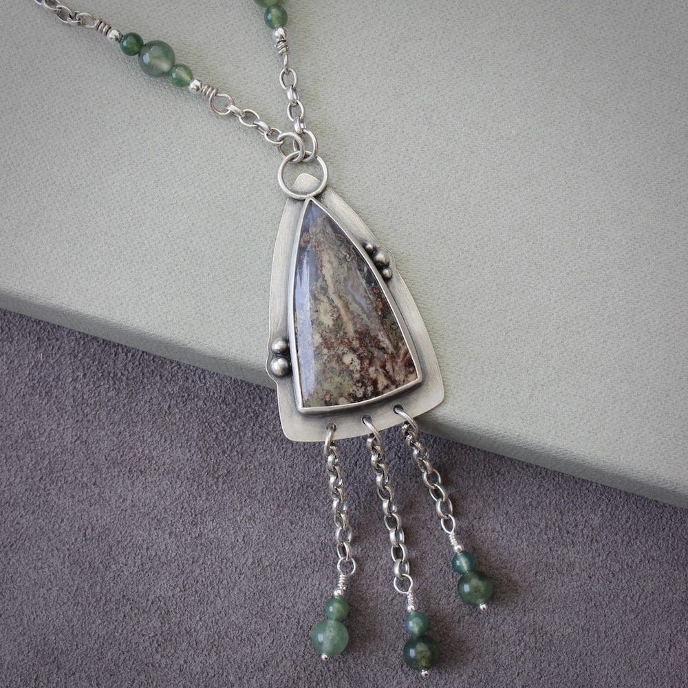 A sterling silver pendant shaped like an arrowhead, with a moss agate gemstone and round moss agate beads on a silver chain. Resting on a green box with a purple suede background.