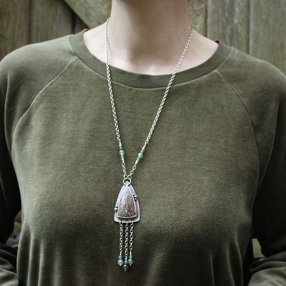 A sterling silver pendant shaped like an arrowhead, with a moss agate gemstone and round moss agate beads on a silver chain, worn by a woman wearing a green velvet jumper standing in front of a wooden gate.