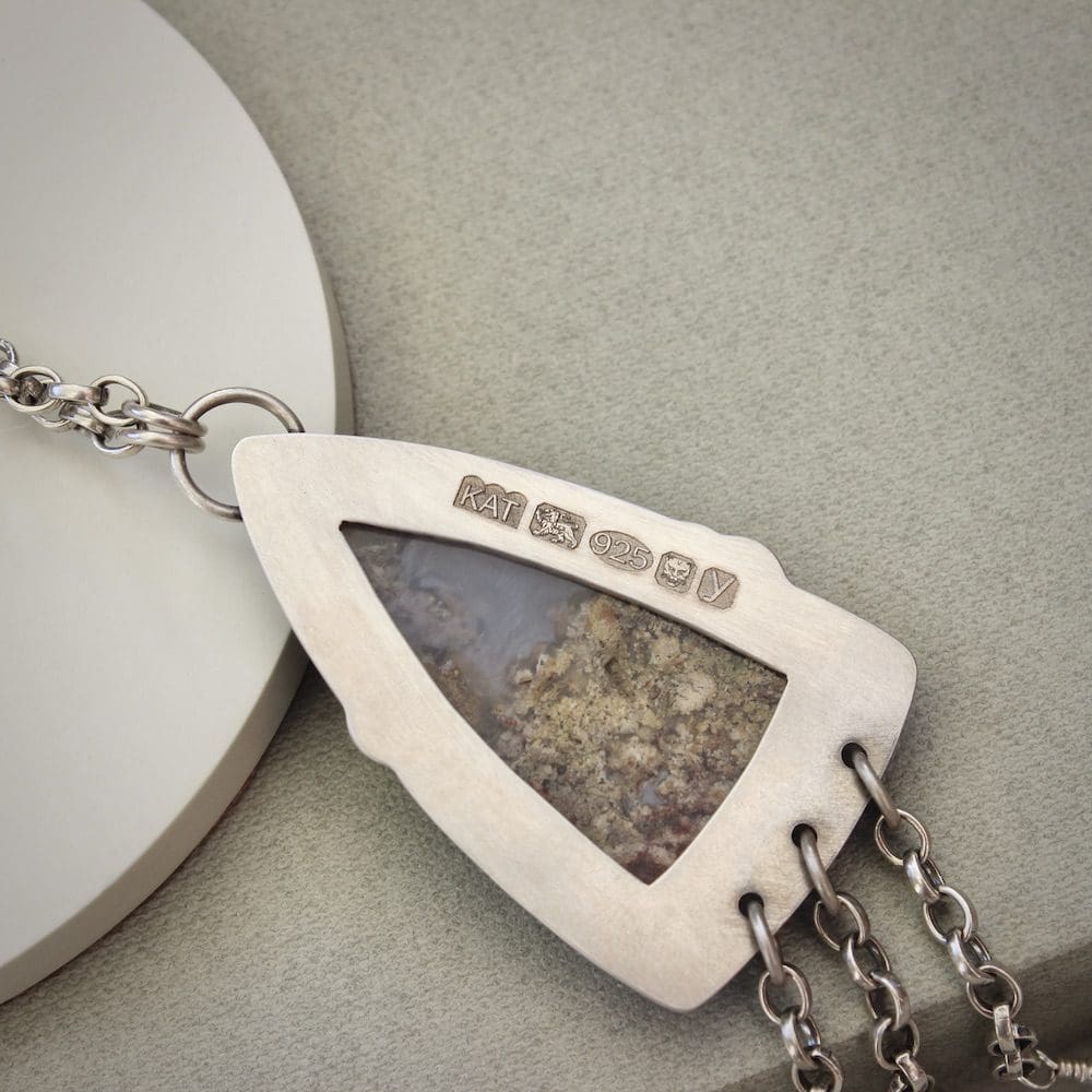 A sterling silver pendant shaped like an arrowhead, with a moss agate gemstone and round moss agate beads on a silver chain. Resting on a green box and shown from the back with a UK hallmark.