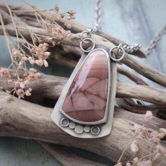 A sterling silver and pink imperial jasper statement pendant on a silver chain, lying on a piece of driftwood against a blue background, framed by tiny pink flowers.