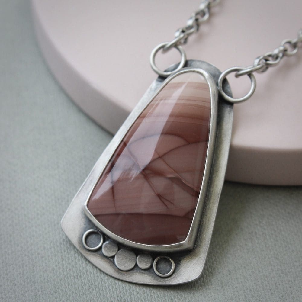 A sterling silver and pink imperial jasper statement pendant on a silver chain, leaning against a circular pink ceramic coaster on a green background.