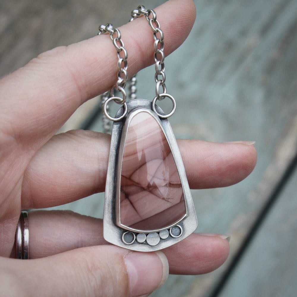 A sterling silver and pink imperial jasper statement pendant on a silver chain, held in the left hand of a woman, above a weathered blue wooden table.