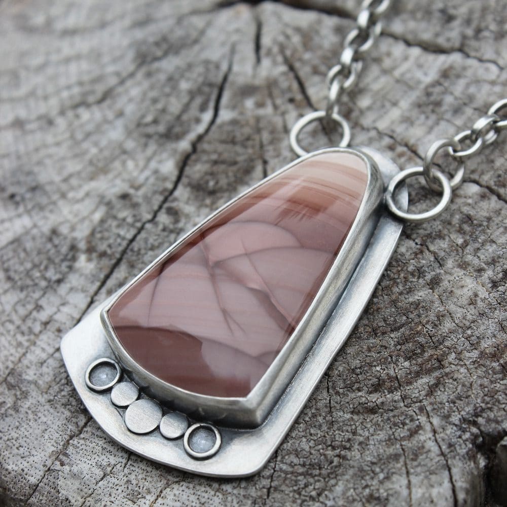 A sterling silver and pink imperial jasper statement pendant on a silver chain, lying on a driftwood log.