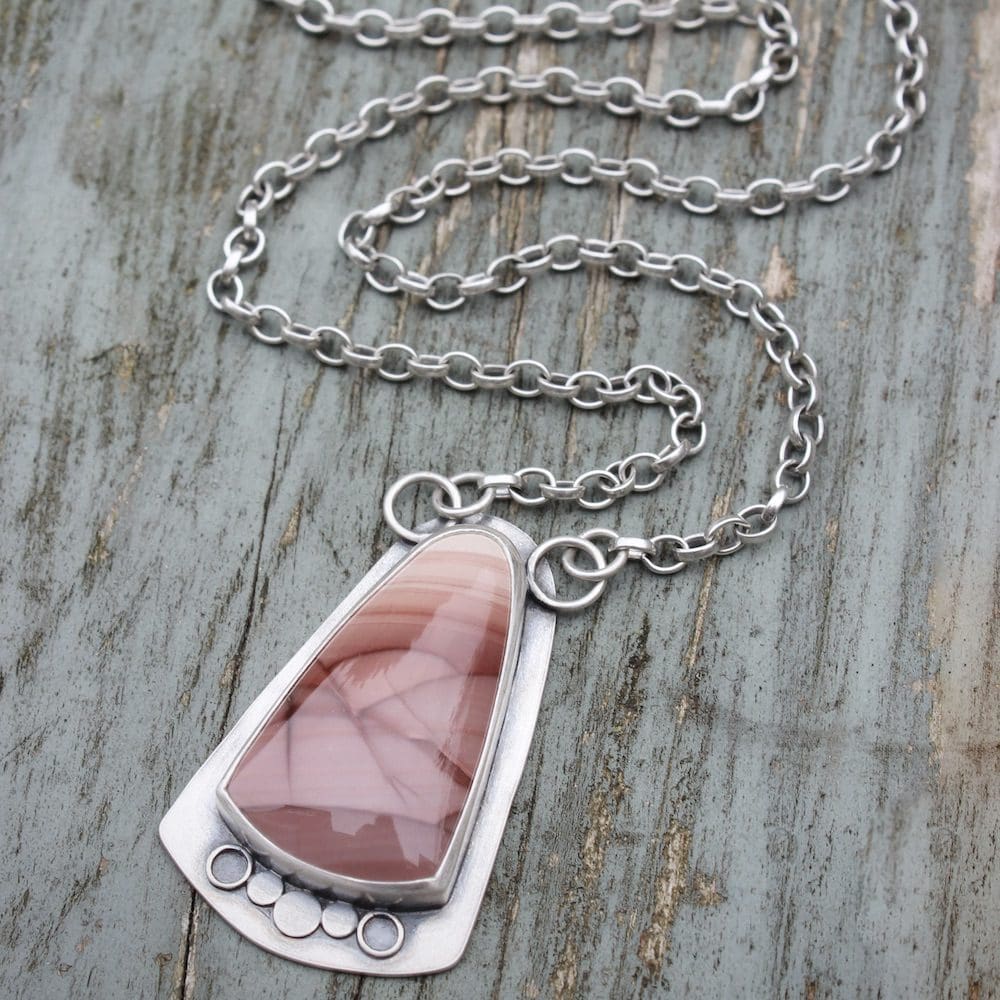 A sterling silver and pink imperial jasper statement pendant on a silver chain, shown laid out in full on a weathered blue wooden background.