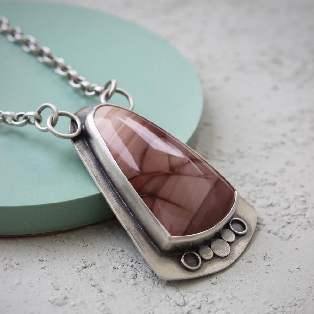 A sterling silver and pink imperial jasper statement pendant on a silver chain, viewed from the side while resting on a green ceramic circular coaster on a pale green background.