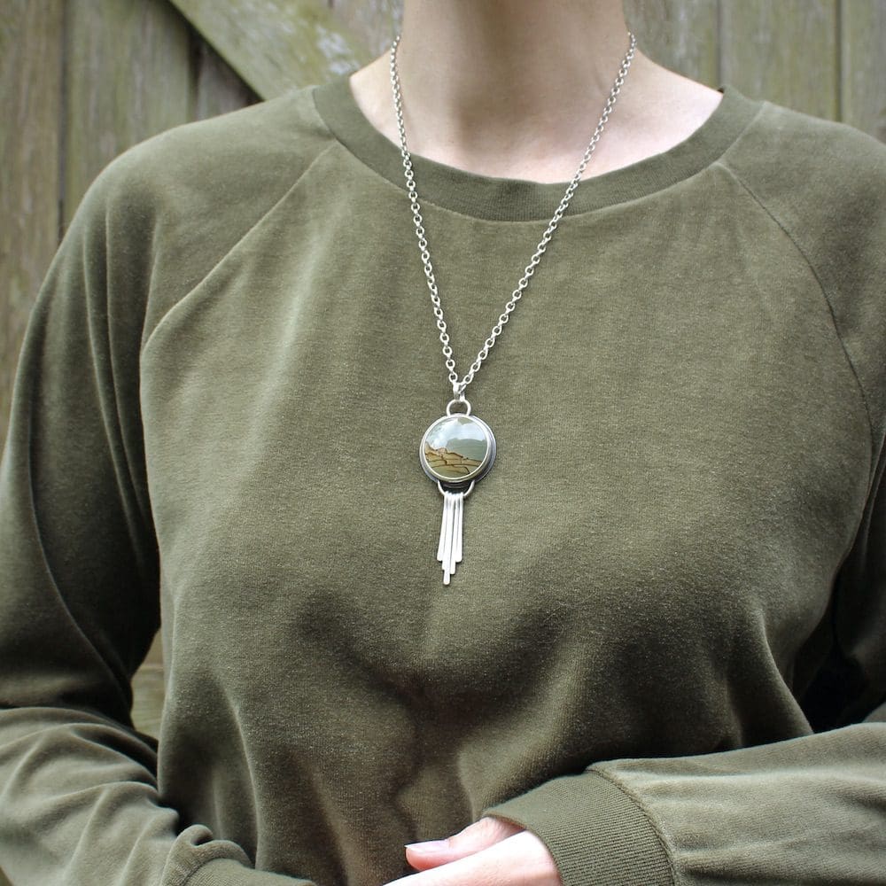 A circular jasper and sterling silver pendant necklace with silver fringing details, worn by a woman wearing a green velvet jumper and standing in front of a wooden gate.