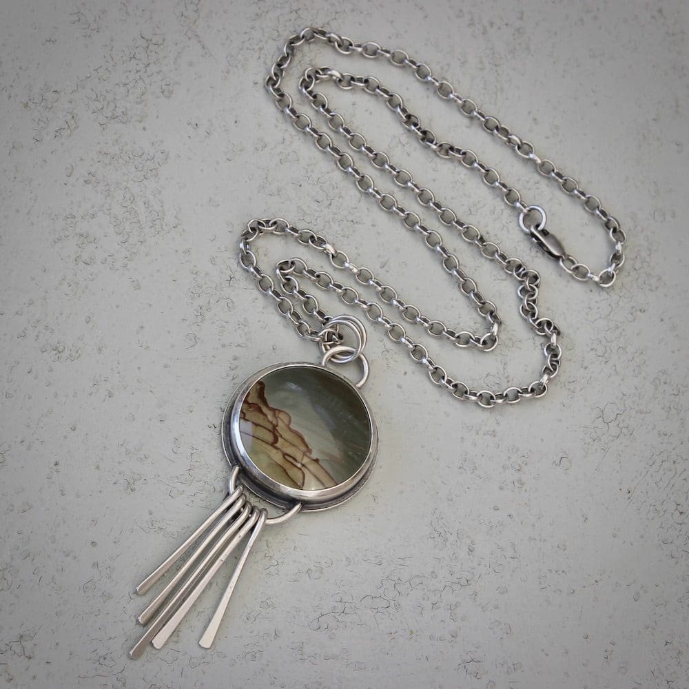 A circular jasper and sterling silver pendant necklace with silver fringing details, lying on a sage green background
