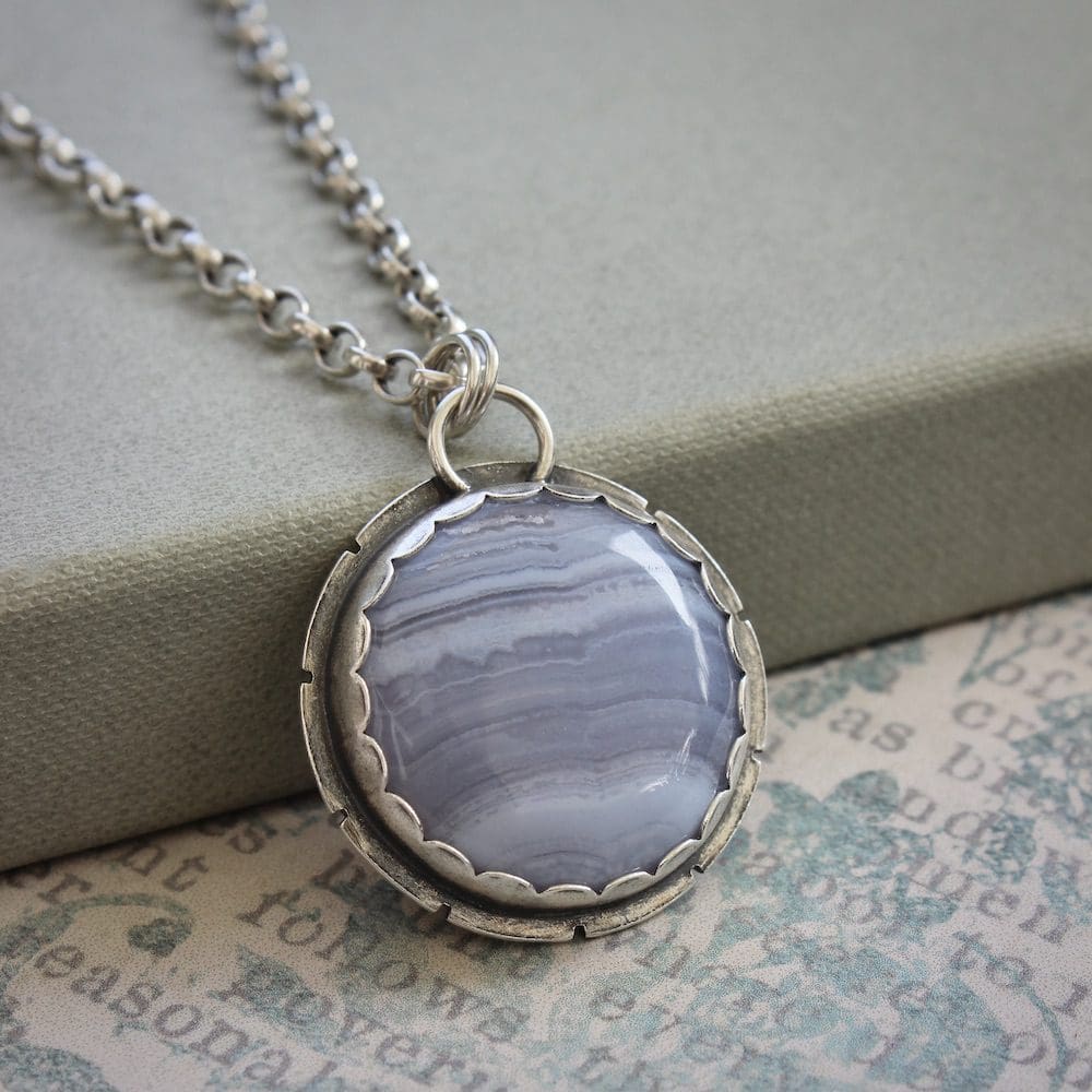 A circular blue lace agate and sterling silver gemstone pendant on a silver chain, propped against a green cardboard box lid on a background with blue and cream flowers and old font text printed on it.