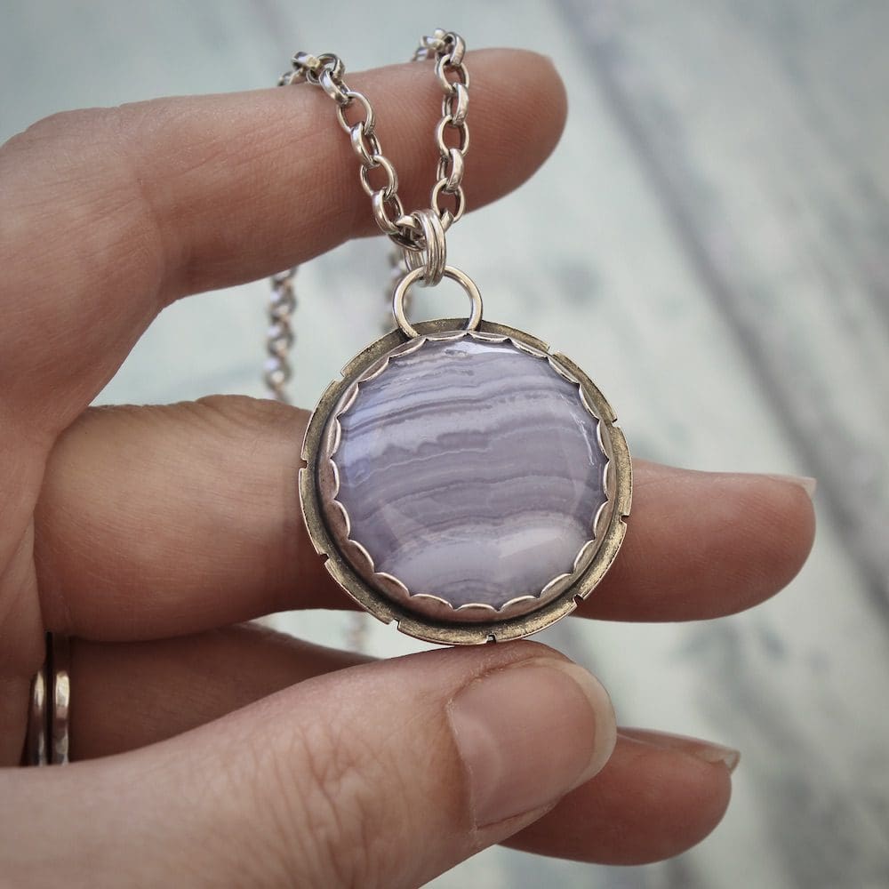 A circular blue lace agate and sterling silver gemstone pendant on a silver chain, held in the left hand of a woman.