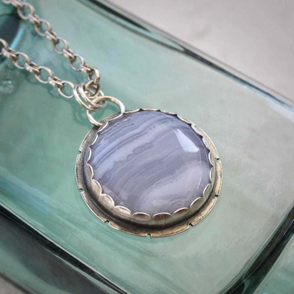 A circular blue lace agate and sterling silver gemstone pendant on a silver chain, lying on an antique green glass bottle.