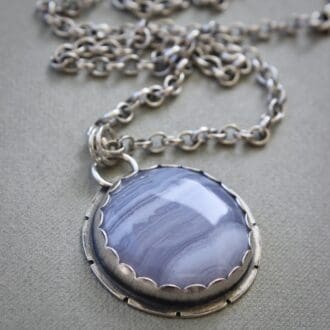 A circular blue lace agate and sterling silver gemstone pendant on a silver chain, on a sage green background.