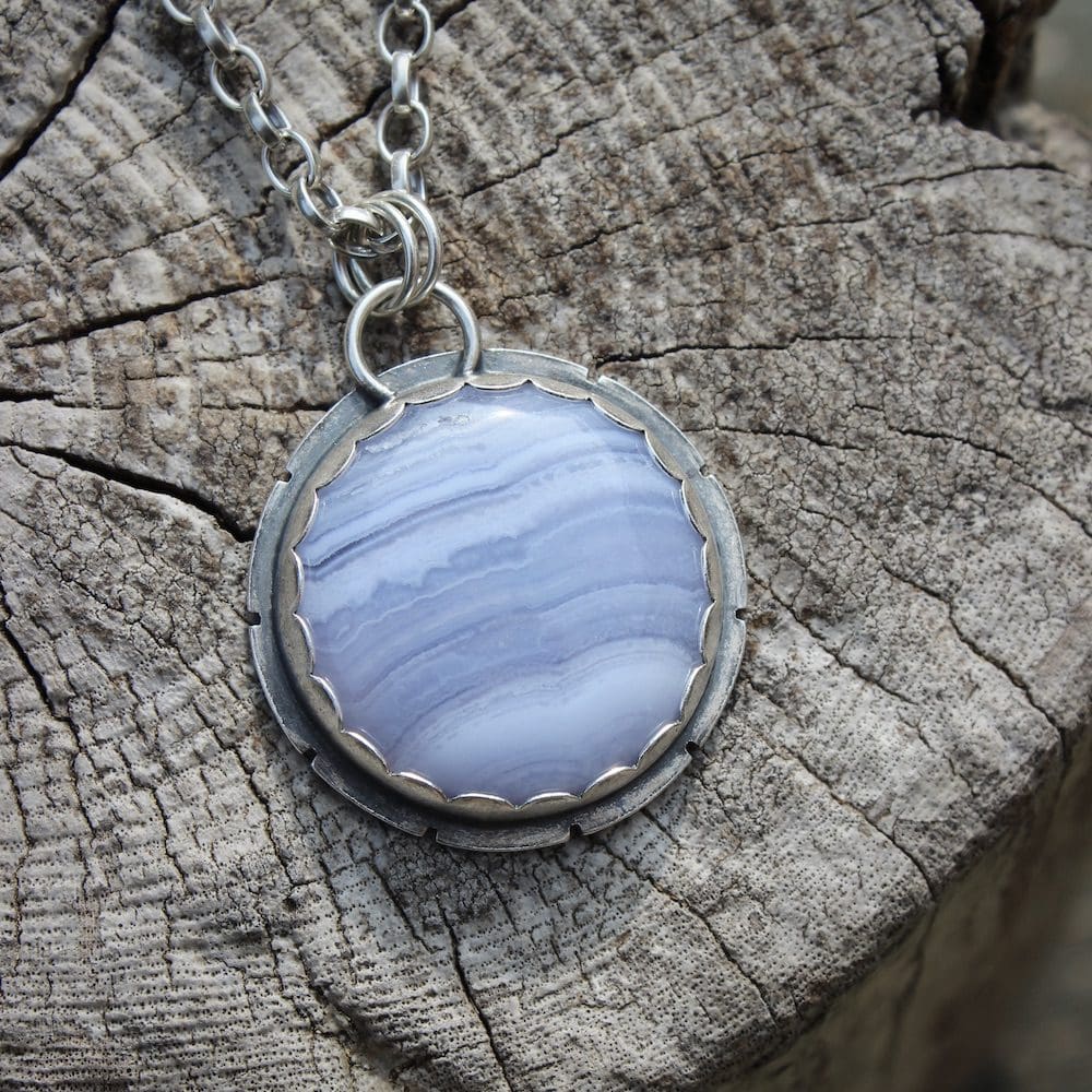 A circular blue lace agate and sterling silver gemstone pendant on a silver chain, sitting on a driftwood log.